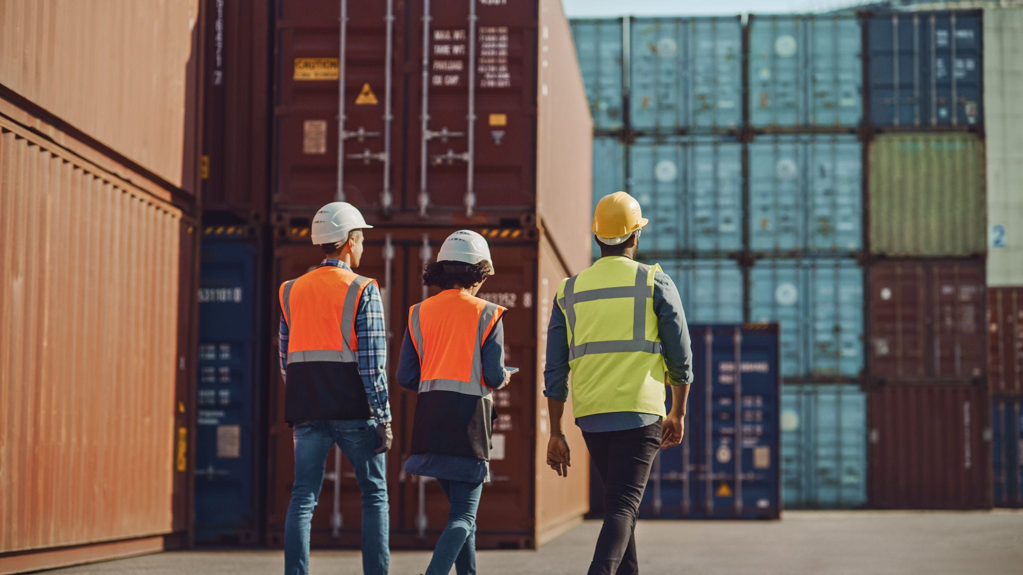 Team of Diverse Industrial Engineers, Safety Supervisors and Foremen in Hard Hats and Safety Vests Walking in Shipping Cargo Container Terminal Depot. Colleagues Talk About Logistics Operations.